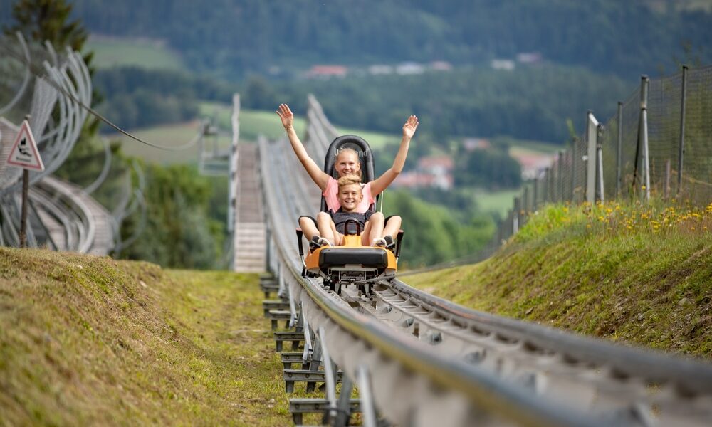 Ein Erwachsener und ein Kind fahren gemeinsam in einem Achterbahnwagen für zwei Personen auf einer von Gras und Hügeln umgebenen Außenbahn.