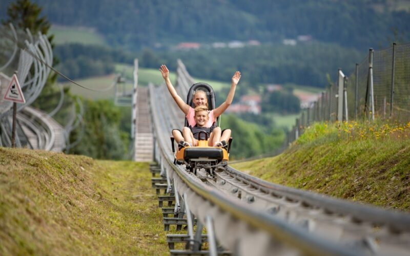 Ein Erwachsener und ein Kind fahren gemeinsam in einem Achterbahnwagen für zwei Personen auf einer von Gras und Hügeln umgebenen Außenbahn.