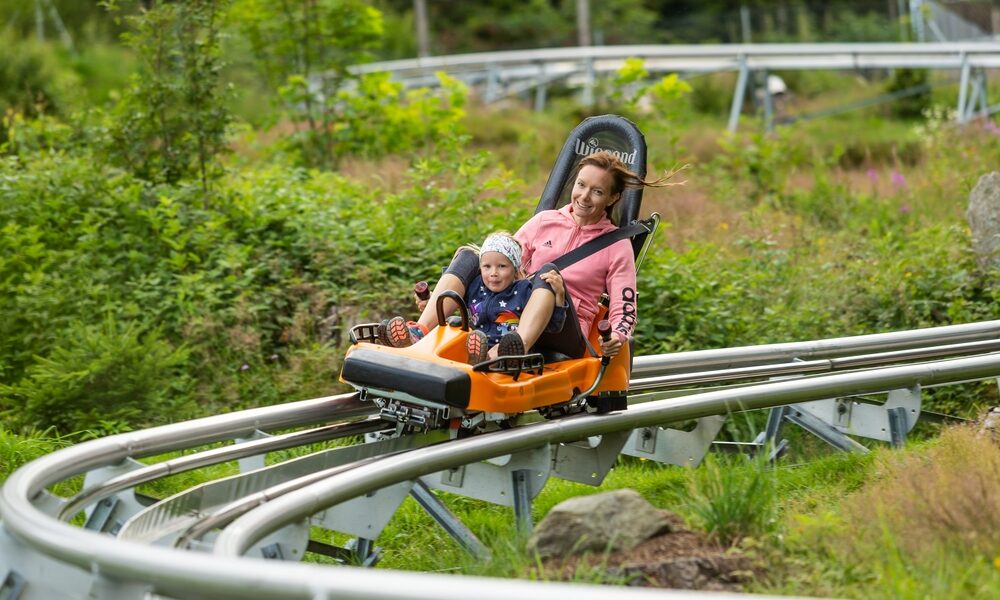 Ein Erwachsener und ein Kind fahren gemeinsam in einem orangefarbenen Alpine-Coaster-Wagen auf einer Metallschiene durch eine grüne, bewaldete Gegend.