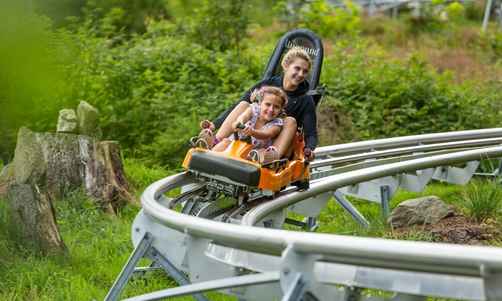 Zwei Personen fahren gemeinsam in einem kleinen orangefarbenen Achterbahnwagen auf einer Metallschiene, umgeben von Grün und Bäumen.