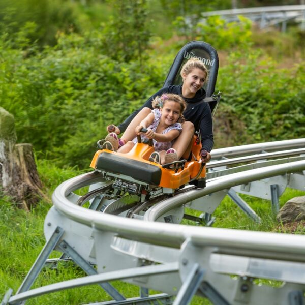 Zwei Personen fahren gemeinsam in einem kleinen orangefarbenen Achterbahnwagen auf einer Metallschiene, umgeben von Grün und Bäumen.