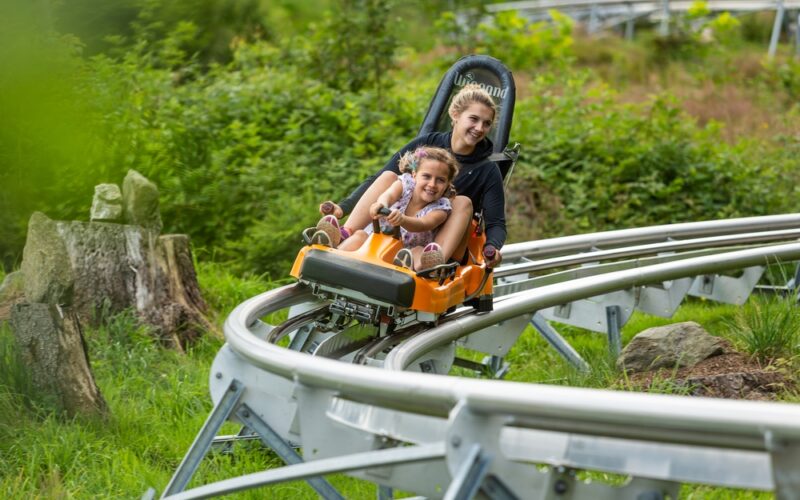 Zwei Personen fahren gemeinsam in einem kleinen orangefarbenen Achterbahnwagen auf einer Metallschiene, umgeben von Grün und Bäumen.