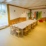 Ein hell erleuchtetes Klassenzimmer mit einem langen Tisch, umgeben von Holzstühlen, bunten Wanddekorationen und einer Spielecke mit kleinen roten Stühlen in der Ecke.