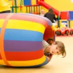 Ein kleines Kind lugt aus einem großen, bunten Spielzylinder in einem Indoor-Spielbereich mit Spielzeugautos und Spielgeräten im Hintergrund hervor.