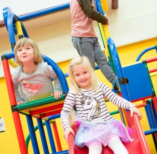 Drei kleine Kinder spielen auf bunten Indoor-Spielgeräten; eines rutscht hinunter, während die beiden anderen klettern und zusehen.