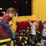 Vier kleine Kinder spielen und werfen Bälle in einer Ballgrube mit gelben und roten Wänden.