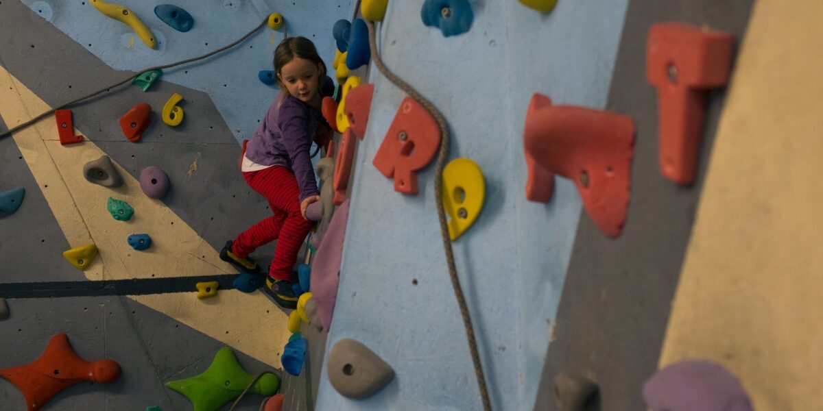 Ein junges Mädchen in lila Jacke und roter Hose klettert an einer Indoor-Kletterwand mit bunten Griffen.