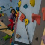 Ein junges Mädchen in lila Jacke und roter Hose klettert an einer Indoor-Kletterwand mit bunten Griffen.