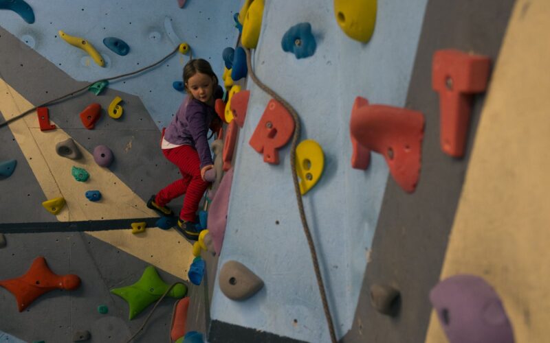 Ein junges Mädchen in lila Jacke und roter Hose klettert an einer Indoor-Kletterwand mit bunten Griffen.