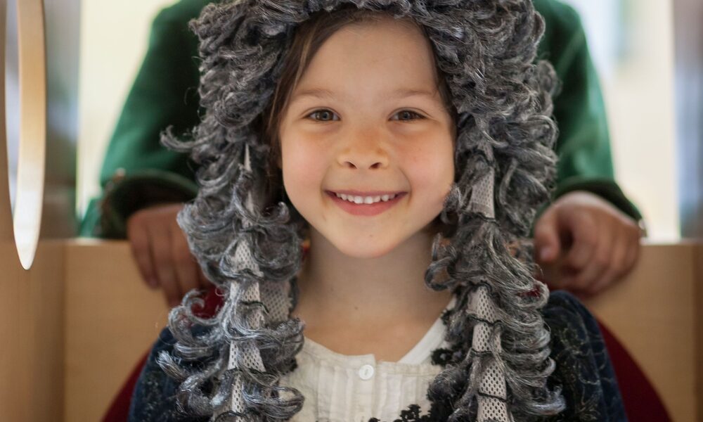 Zwei Kinder in historischen Kostümen; das Mädchen vorne trägt eine lockige graue Perücke und ein rot-schwarzes Kleid, während das Kind hinten einen Dreispitz und einen grünen Mantel trägt.
