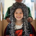 Zwei Kinder in historischen Kostümen; das Mädchen vorne trägt eine lockige graue Perücke und ein rot-schwarzes Kleid, während das Kind hinten einen Dreispitz und einen grünen Mantel trägt.