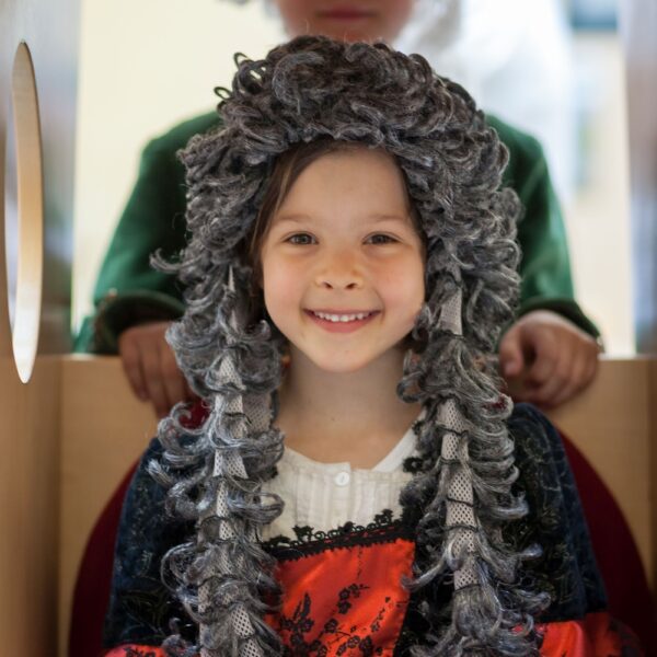 Zwei Kinder in historischen Kostümen; das Mädchen vorne trägt eine lockige graue Perücke und ein rot-schwarzes Kleid, während das Kind hinten einen Dreispitz und einen grünen Mantel trägt.