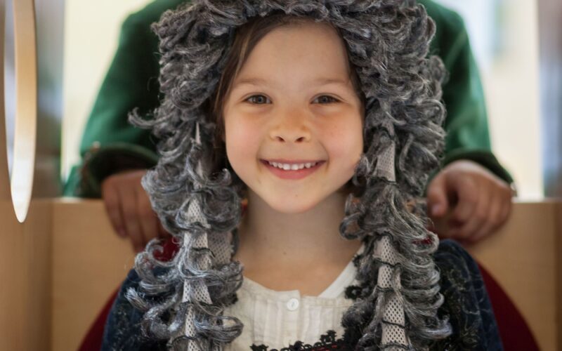 Zwei Kinder in historischen Kostümen; das Mädchen vorne trägt eine lockige graue Perücke und ein rot-schwarzes Kleid, während das Kind hinten einen Dreispitz und einen grünen Mantel trägt.