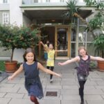 Drei kleine Kinder rennen und lächeln vor einem Gebäudeeingang, umgeben von Topfpflanzen und Grünpflanzen auf einem gepflasterten Gehweg.