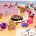 Ein Geburtstagstisch ist mit einem Schokoladenkuchen, bunten Kronen, Fotos, Federschmuck und einem Schloss aus rosa Karton gedeckt.