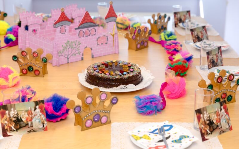 Ein Geburtstagstisch ist mit einem Schokoladenkuchen, bunten Kronen, Fotos, Federschmuck und einem Schloss aus rosa Karton gedeckt.