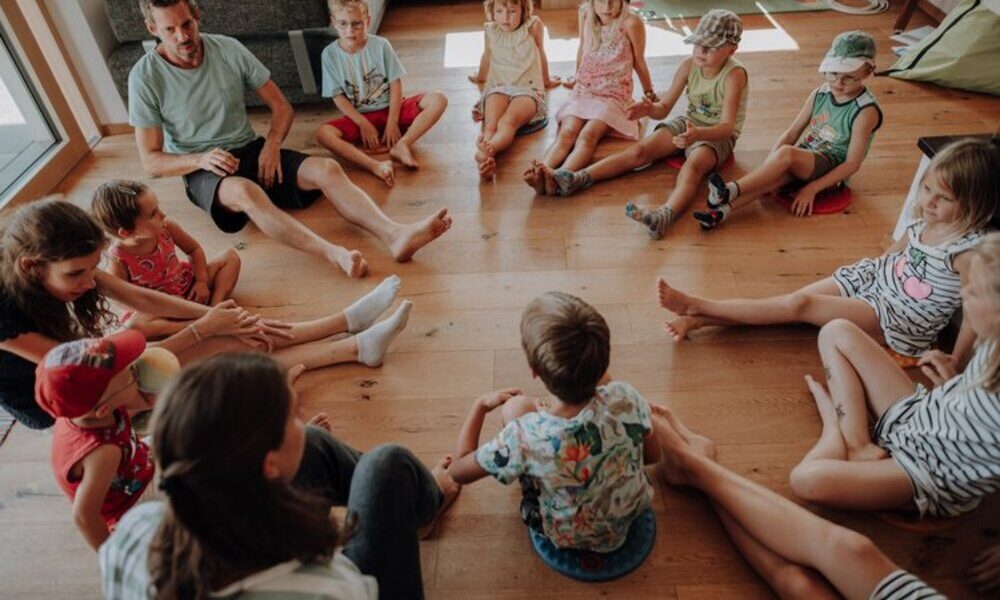 Eine Gruppe von Kindern und zwei Erwachsene sitzen im Kreis auf einem Holzboden und nehmen an einer Gruppenaktivität oder einer Diskussion in den Innenräumen teil.