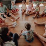 Eine Gruppe von Kindern und zwei Erwachsene sitzen im Kreis auf einem Holzboden und nehmen an einer Gruppenaktivität oder einer Diskussion in den Innenräumen teil.
