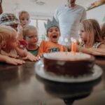 Eine Gruppe von Kindern versammelt sich um eine Geburtstagstorte mit brennenden Kerzen; ein Kind mit einer Papierkrone sitzt aufgeregt in der Mitte.