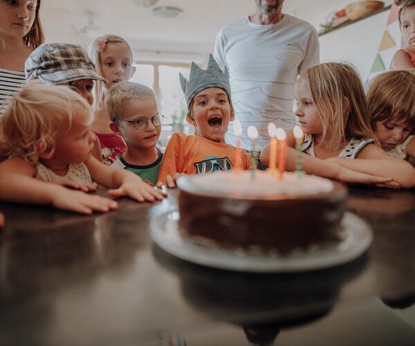 Eine Gruppe von Kindern versammelt sich um eine Geburtstagstorte mit brennenden Kerzen; ein Kind mit einer Papierkrone sitzt aufgeregt in der Mitte.