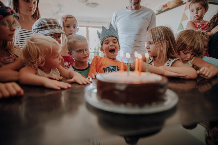 Eine Gruppe von Kindern versammelt sich um eine Geburtstagstorte mit brennenden Kerzen; ein Kind mit einer Papierkrone sitzt aufgeregt in der Mitte.