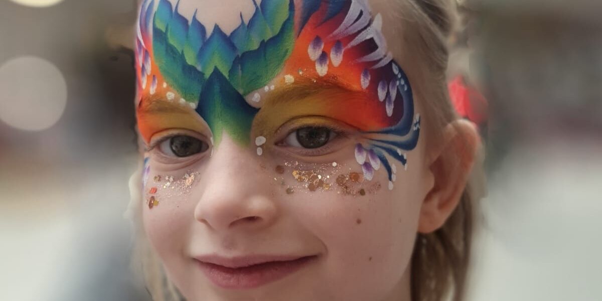 Kleinkind mit bunter, aufwendiger Gesichtsbemalung, die einem Schmetterling oder einer Maske ähnelt, mit Glitzerakzenten und einem violetten Oberteil; unscharfer Hintergrund in einem Innenraum.