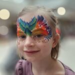 Kleinkind mit bunter, aufwendiger Gesichtsbemalung, die einem Schmetterling oder einer Maske ähnelt, mit Glitzerakzenten und einem violetten Oberteil; unscharfer Hintergrund in einem Innenraum.