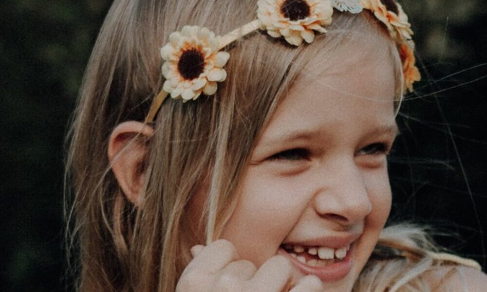 Ein junges Mädchen mit langen blonden Haaren trägt ein Sonnenblumen-Stirnband und lächelt, während es zur Seite blickt; im Hintergrund ist Grün zu sehen.