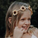 Ein junges Mädchen mit langen blonden Haaren trägt ein Sonnenblumen-Stirnband und lächelt, während es zur Seite blickt; im Hintergrund ist Grün zu sehen.