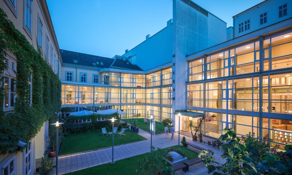 Ein modernes Gebäude mit Glaswänden umgibt einen Innenhof mit Bänken, Tischen und Grünflächen, die abends von der Innenbeleuchtung erhellt werden.
