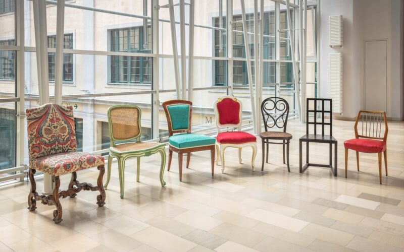 Sechs verschiedene Vintage-Stühle stehen in einer Reihe auf einem gefliesten Boden vor großen Fenstern mit Metallrahmen.