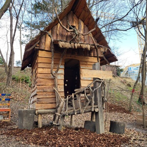 Ein kleines hölzernes Spielhaus mit steilem Dach, umgeben von Bäumen und Mulch, verfügt über ein rustikales Astgeländer am Eingang.