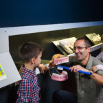 Ein Erwachsener kniet neben zwei Kindern und demonstriert das Zähneputzen an einem großen Zahnmodell mit einer riesigen Zahnbürste in einer Museumsausstellung.