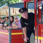 Ein lächelnder Zugführer lehnt sich aus einem roten Touristenzug mit der Aufschrift "Untersberg", während Fahrgäste, darunter Erwachsene und Kinder, hinausschauen und winken. Im Hintergrund sind Bäume zu sehen.