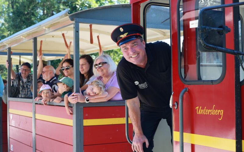 Ein lächelnder Zugführer lehnt sich aus einem roten Touristenzug mit der Aufschrift "Untersberg", während Fahrgäste, darunter Erwachsene und Kinder, hinausschauen und winken. Im Hintergrund sind Bäume zu sehen.