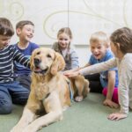 Fünf Kinder sitzen in einem Kreis auf dem Boden und streicheln einen großen Golden Retriever. Die Kinder wirken glücklich und beschäftigt mit dem Hund.
