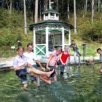 Eine Gruppe von Kindern und Erwachsenen steht und spielt in einem flachen Außenbecken in der Nähe eines kleinen weißen Pavillons, umgeben von Bäumen und Grün.