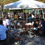 An einem sonnigen Tag sitzen Menschen an Holztischen mit Metallstühlen in einem Café im Freien, das von großen weißen Sonnenschirmen beschattet wird.