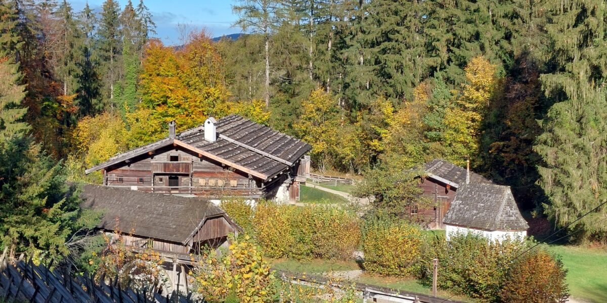 Holzhäuser mit Schindeldächern schmiegen sich zwischen herbstlich belaubte Bäume, die von einem rustikalen Holzzaun begrenzt werden, unter einem teilweise bewölkten Himmel.