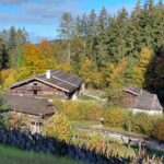 Holzhäuser mit Schindeldächern schmiegen sich zwischen herbstlich belaubte Bäume, die von einem rustikalen Holzzaun begrenzt werden, unter einem teilweise bewölkten Himmel.