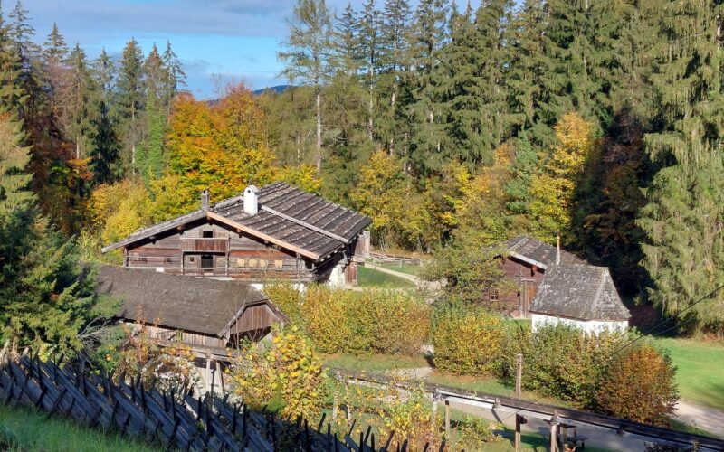Holzhäuser mit Schindeldächern schmiegen sich zwischen herbstlich belaubte Bäume, die von einem rustikalen Holzzaun begrenzt werden, unter einem teilweise bewölkten Himmel.