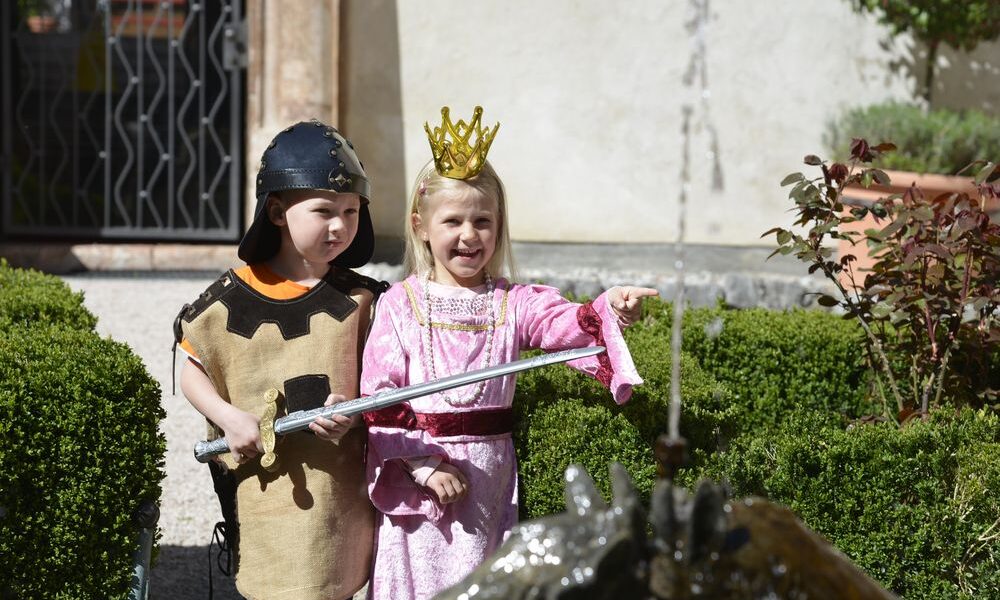Ein Junge, der als Ritter gekleidet ist, und ein Mädchen, das als Prinzessin gekleidet ist, stehen an einem Gartenbrunnen; das Mädchen zeigt auf etwas, während der Junge ein Spielzeugschwert hält.