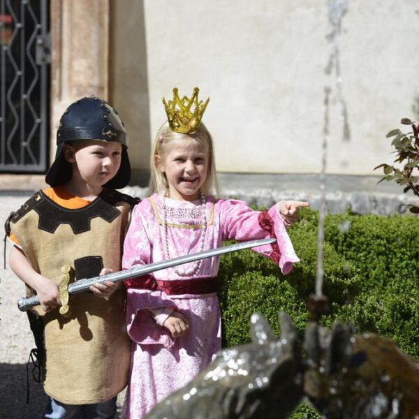 Ein Junge, der als Ritter gekleidet ist, und ein Mädchen, das als Prinzessin gekleidet ist, stehen an einem Gartenbrunnen; das Mädchen zeigt auf etwas, während der Junge ein Spielzeugschwert hält.