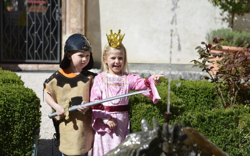 Ein Junge, der als Ritter gekleidet ist, und ein Mädchen, das als Prinzessin gekleidet ist, stehen an einem Gartenbrunnen; das Mädchen zeigt auf etwas, während der Junge ein Spielzeugschwert hält.