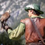 Ein Mann in grüner und brauner Kleidung im mittelalterlichen Stil hält einen Falken auf seiner behandschuhten Hand, im Hintergrund sind verschwommene Berge zu sehen.
