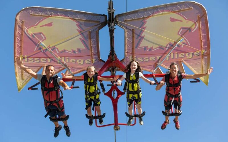 Vier Personen in Klettergurten schweben auf einer Seilrutsche mit großen Adlerflügeln vor einem strahlend blauen Himmel durch die Luft.
