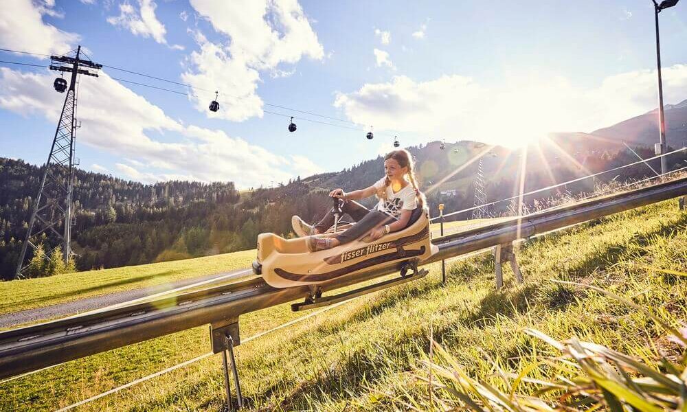 Zwei Personen fahren mit einem Alpine Coaster einen grasbewachsenen Hang hinunter, mit Bäumen, Seilbahnen und Bergen im Hintergrund in der strahlenden Sonne.