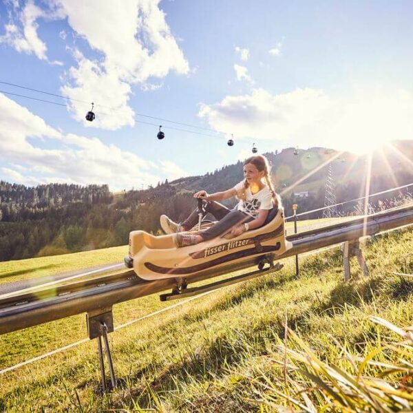 Zwei Personen fahren mit einem Alpine Coaster einen grasbewachsenen Hang hinunter, mit Bäumen, Seilbahnen und Bergen im Hintergrund in der strahlenden Sonne.