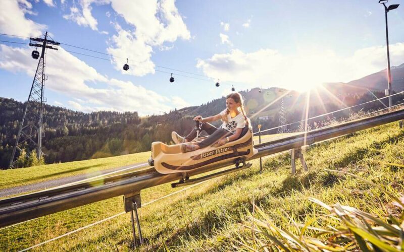 Zwei Personen fahren mit einem Alpine Coaster einen grasbewachsenen Hang hinunter, mit Bäumen, Seilbahnen und Bergen im Hintergrund in der strahlenden Sonne.