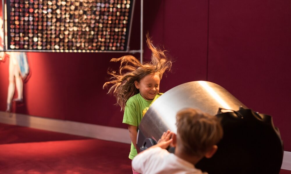 Zwei Kinder spielen zusammen im Haus; ein Kind schiebt einen großen zylindrischen Gegenstand, wodurch sich die Haare des anderen in die Luft heben.
